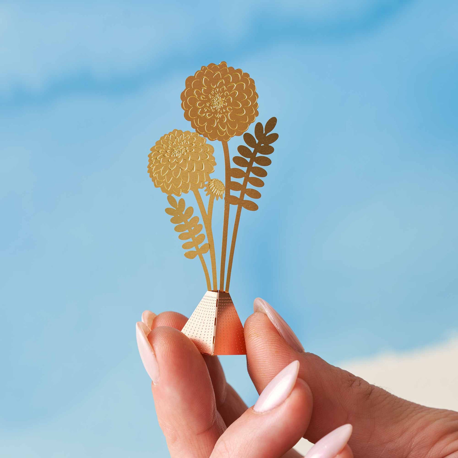 A hand holding a small dahlia bouquet decoration made of brass with a gold finish, with a base and three-dimensional flowers, against a blue background.