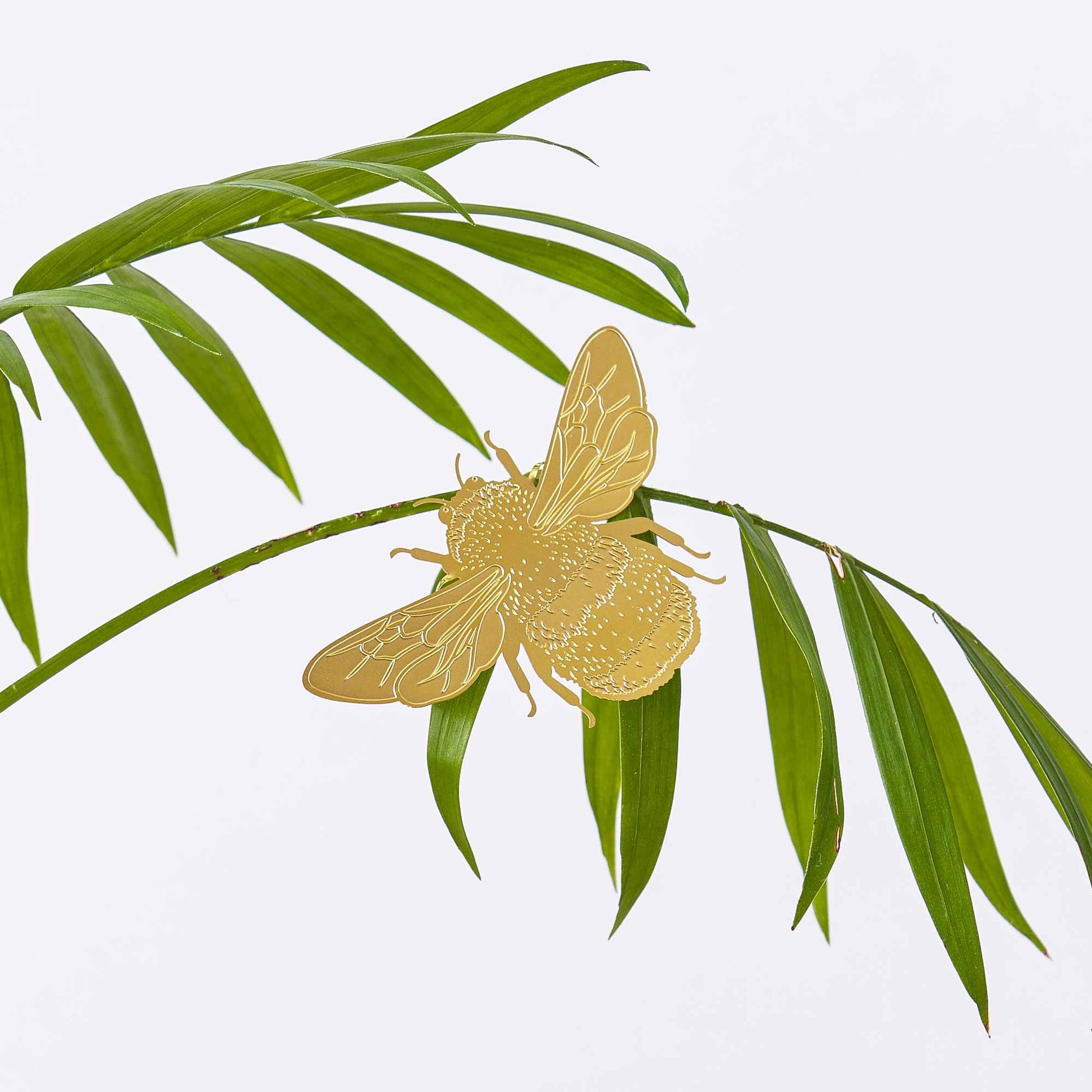 A brass etched bumblebee plant decoration clipped onto a plant, with a beige background and green leaves.