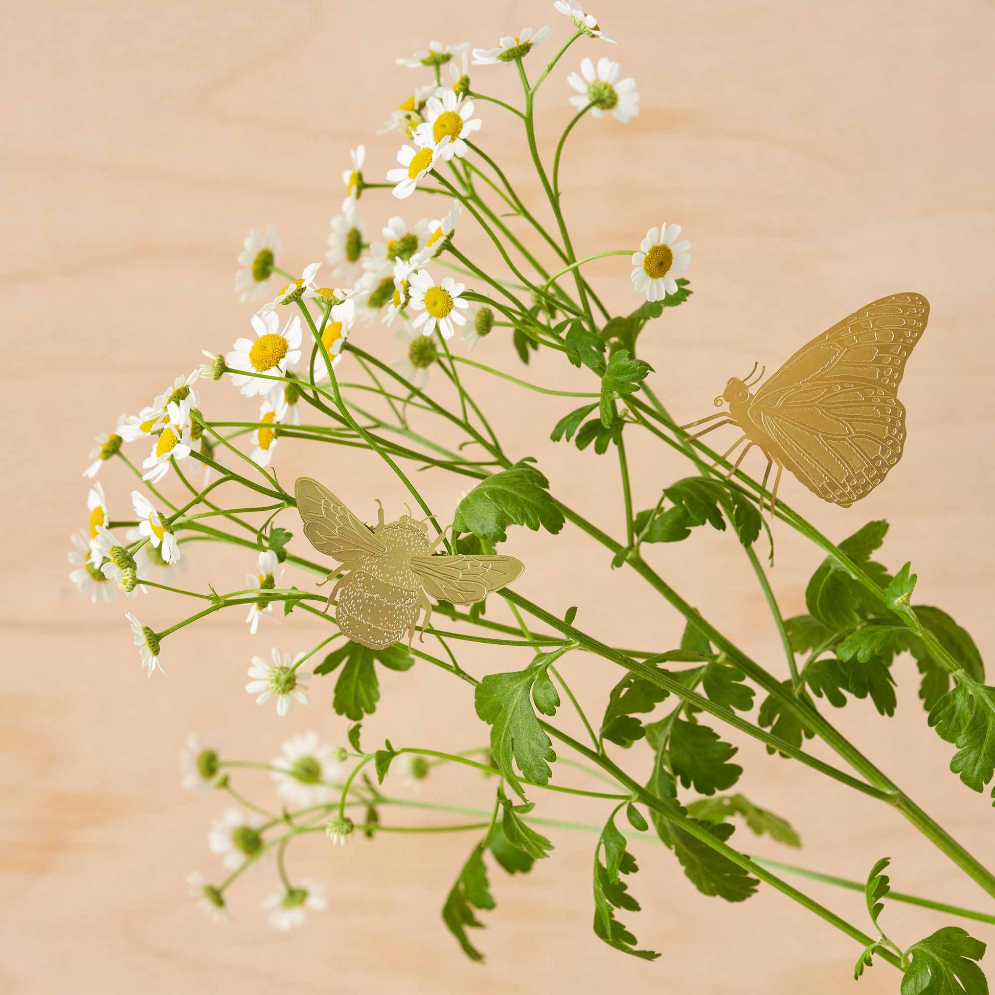 A brass etched bumblebee plant decoration clipped onto a plant, with a beige background and green leaves.