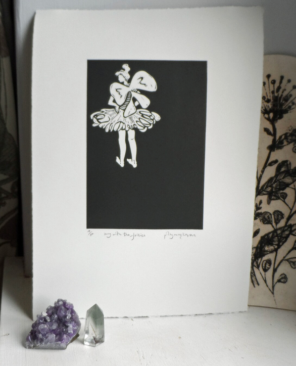 A signed and titled original linocut print by Polly Marix-Evans, depicting a fairy, mounted on white paper. The artwork is displayed with a crystal and a floral patterned object in the background.