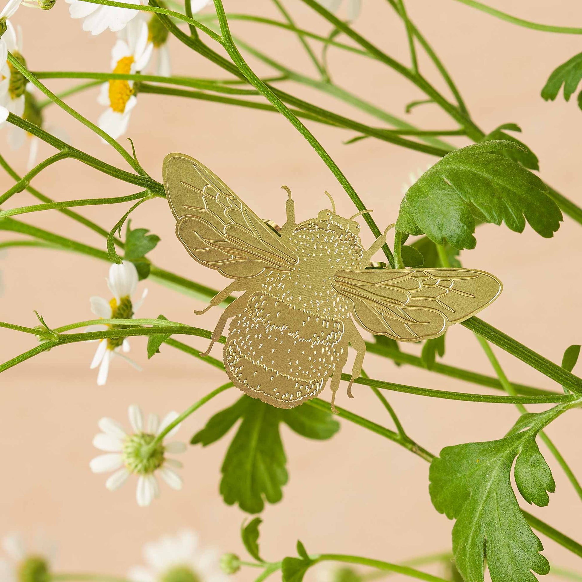 A brass etched bumblebee plant decoration clipped onto a plant, with a beige background and green leaves.