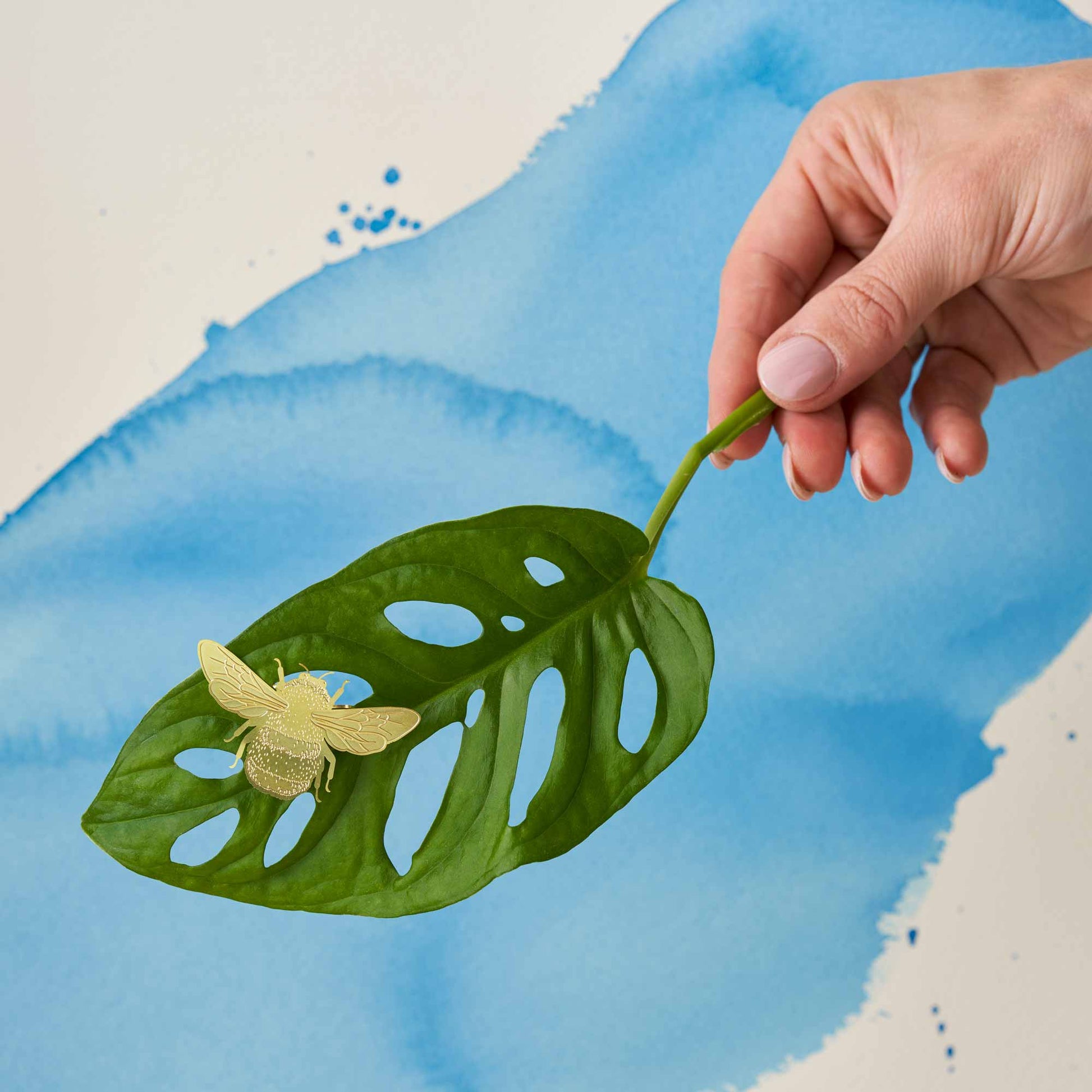 A brass etched bumblebee plant decoration clipped onto a plant, with a blue background and green leaves.