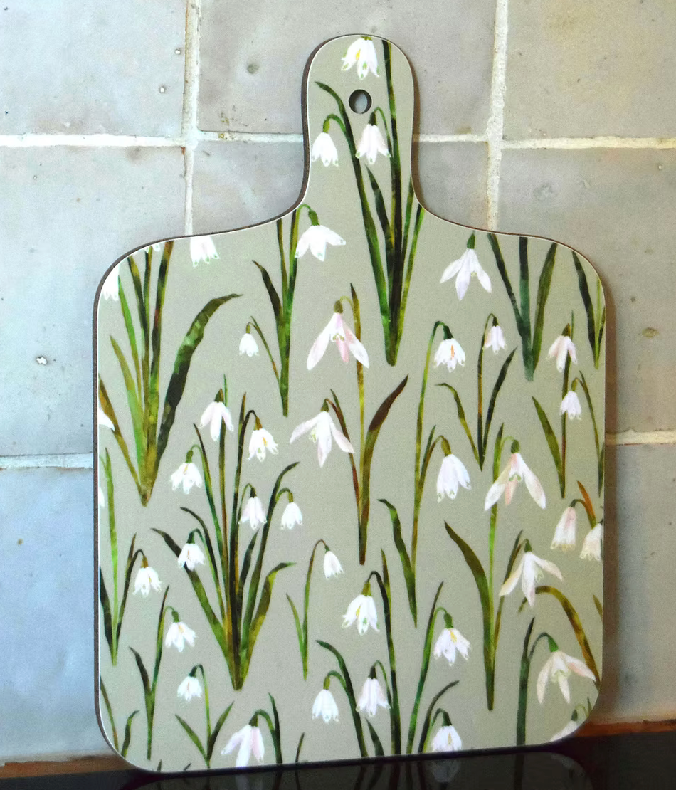 A decorative mini chopping board with a lovely snowdropdesign on a white background, displayed on a wooden surface with vegetables around it.