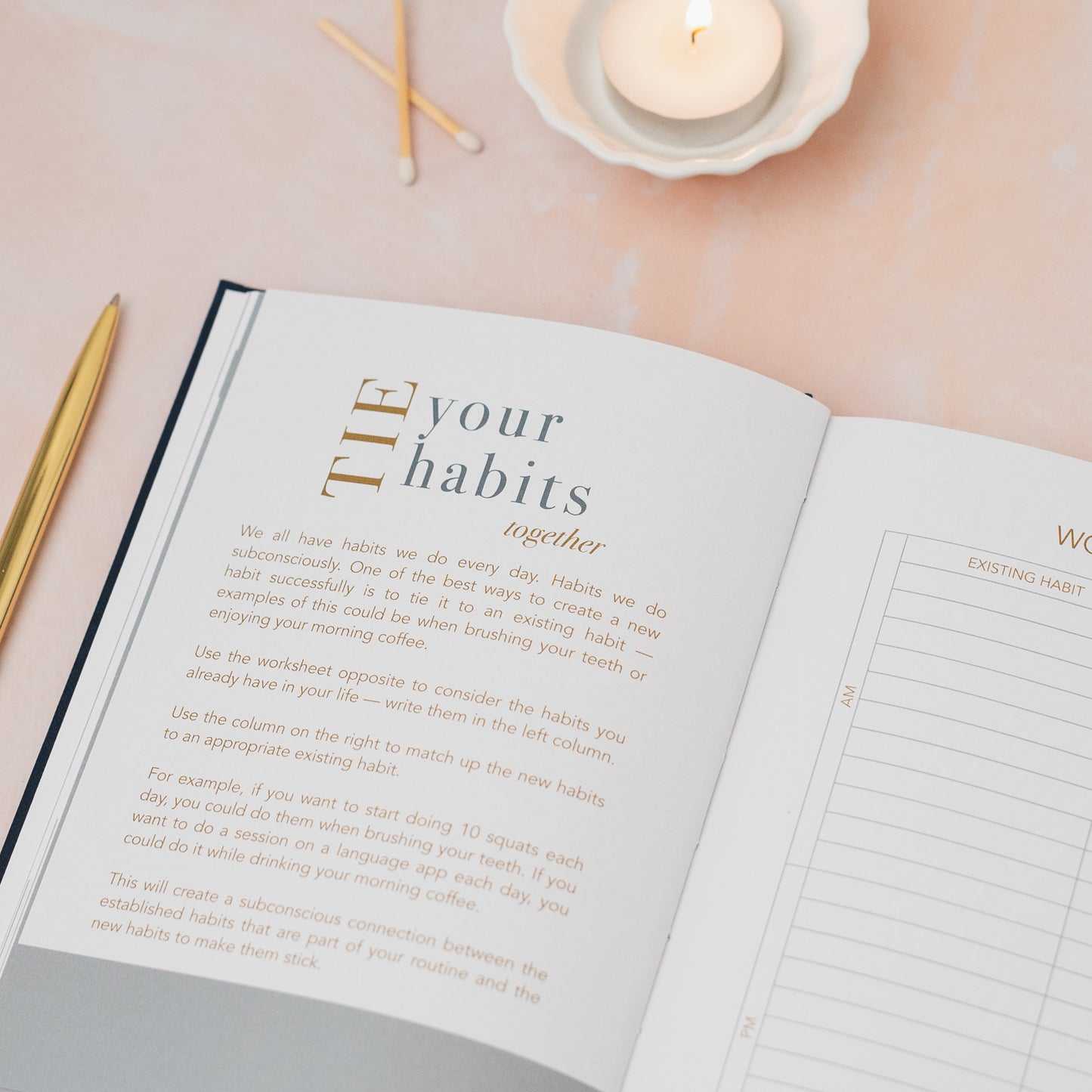 Open book with habit tracker on a soft pink surface with candles and pens.