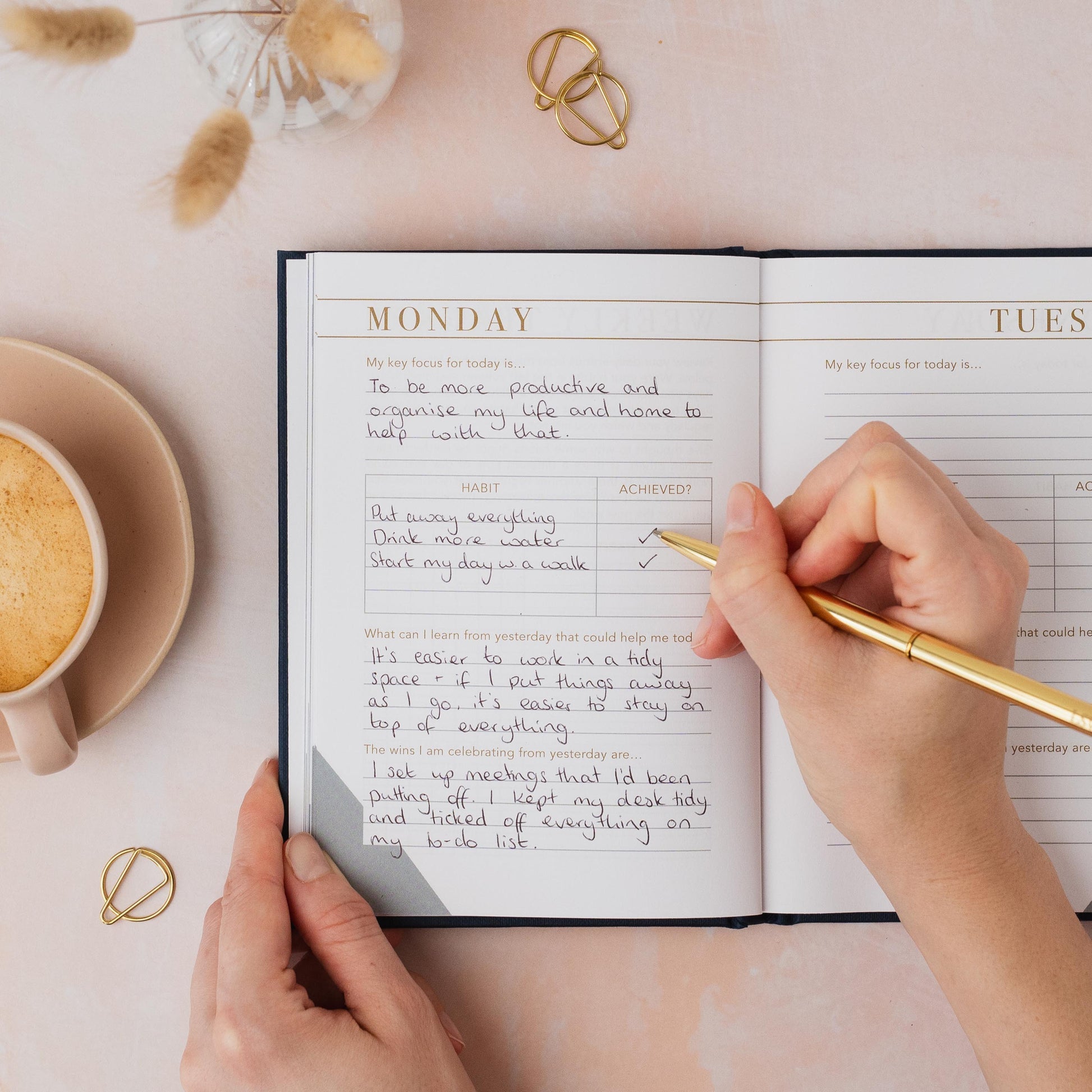 Person writing in a planner with a cup of coffee and decorative items on a table. LSW Habit notes for Heart Gallery