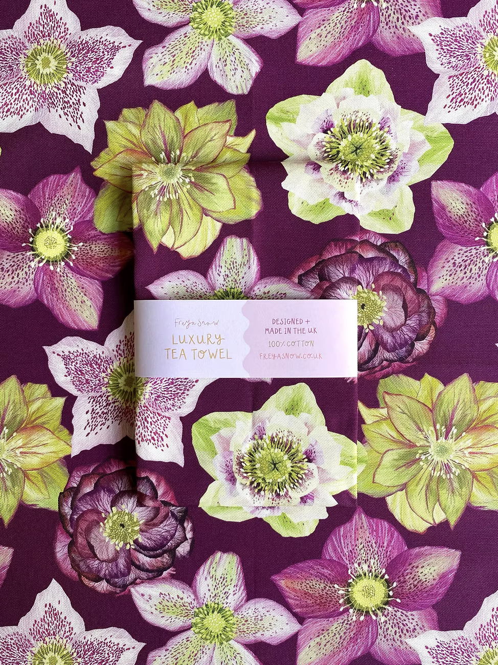 A multicolored floral tea towel with a prominent purple hue and green leaves, featuring a label with the brand name 'Freya Snow'.