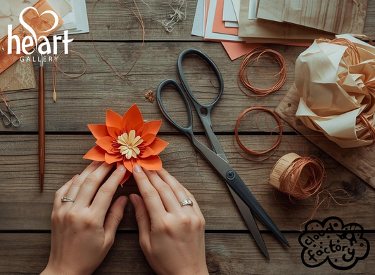 Hands crafting an orange flower with scissors and string on a wooden surface, with 'Heart Gallery' branding and Cloud Factory logo Hebden Bridge