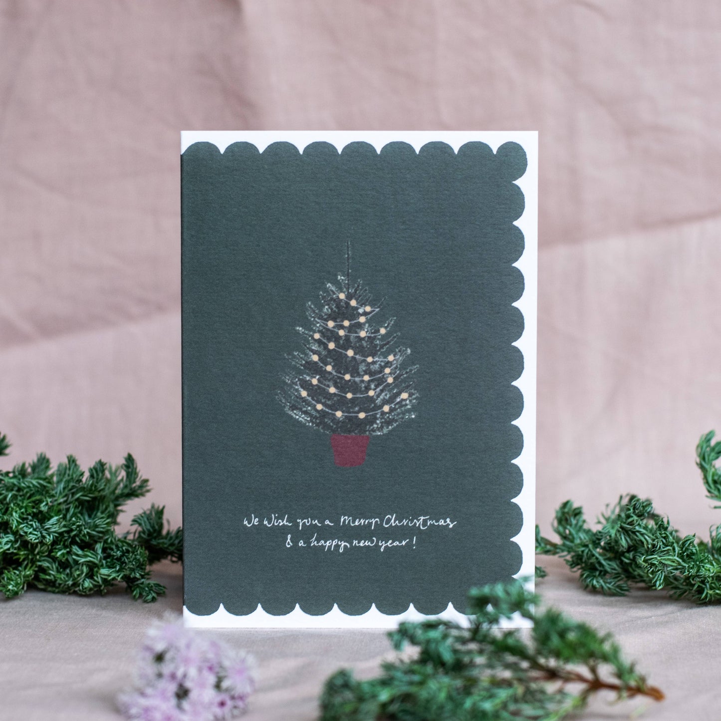 Christmas card with a tree design and text on a scalloped edge, surrounded by greenery.