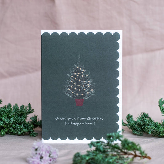 Christmas card with a tree design and text on a scalloped edge, surrounded by greenery.