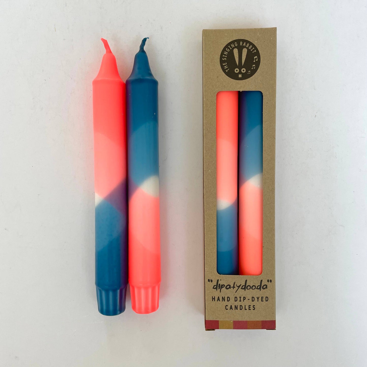 Two hand-dip-dyed candles in neon coral and teal blue with their packaging on a white background by The Singing Rabbit