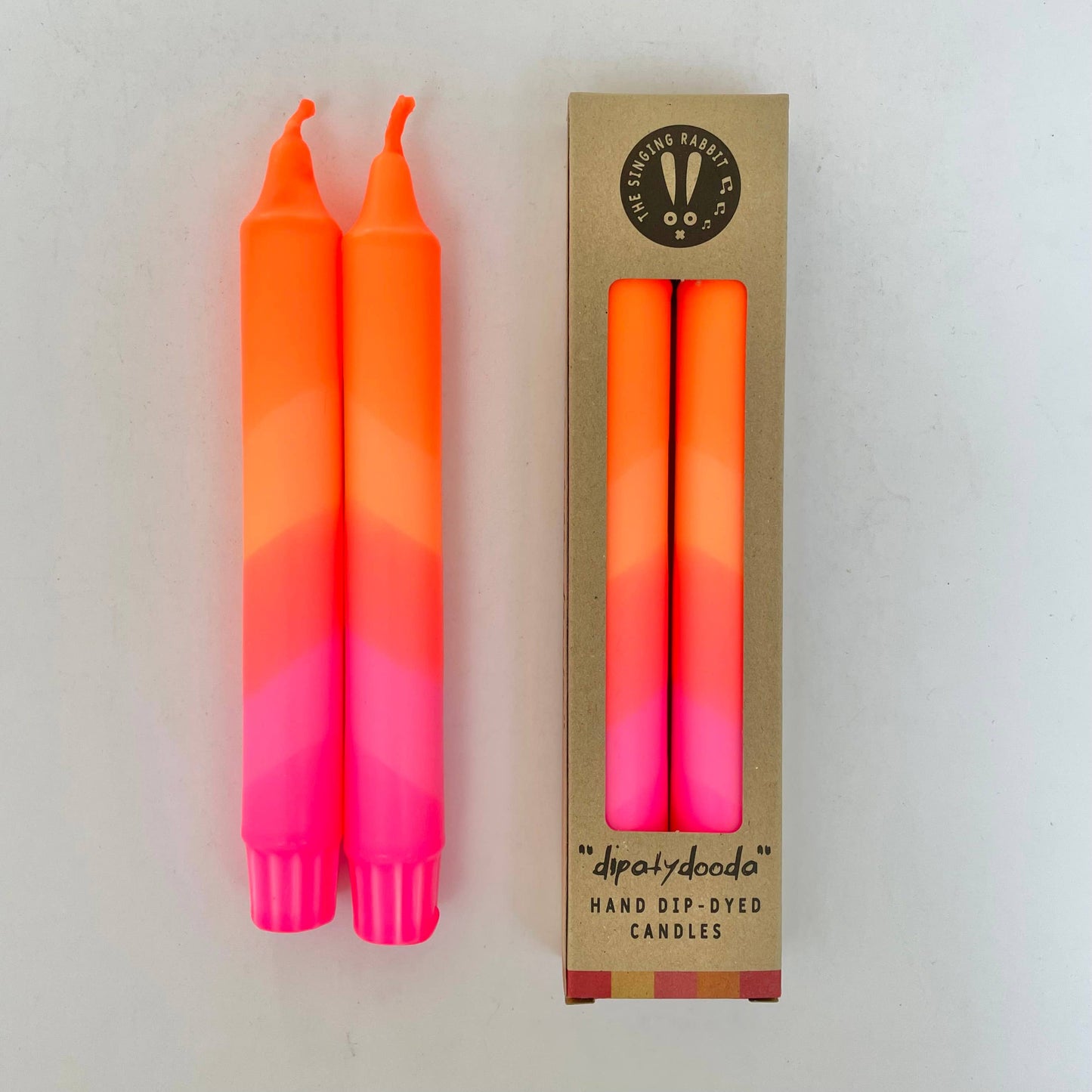 Two dip-dyed candles with a gradient from pink to orange, next to their cardboard packaging by The Singing Rabbit