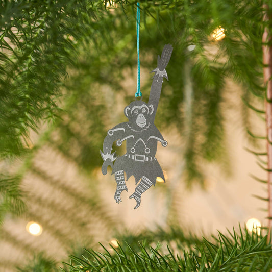 A stainless steel chimpanzee-shaped Christmas ornament hanging from a blue cord, displayed against a plant with a Christmas Tree in the background by Another Studio for Heart Gallery