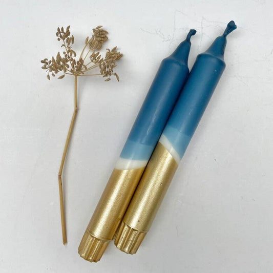 Two dinner candles with a dip dyed design in teal blue and gold, alongside a dried flower decoration.