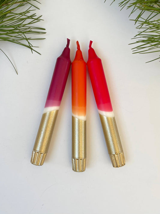 Three candles dip dyed in berry colours with gold bases and colorful wicks on a light background