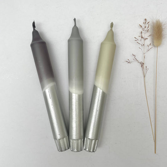 Three dinner candles with a dip-dye design, in shades of grey and silver, displayed alongside some decorative dried plants.