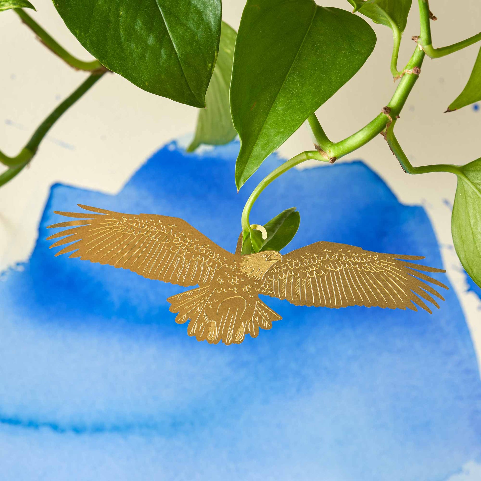 A gold-colored brass plant animal in the shape of an eagle, with open wings, positioned among green plant leaves by Another Studio for Heart Gallery