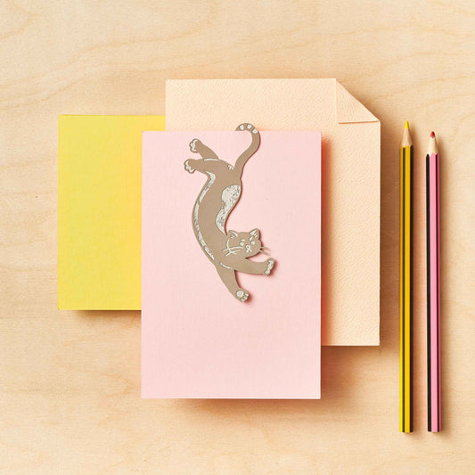 A stainless steel bookmark with an etched cat design, displayed on a pink notepad, with a yellow envelope and pencils in the background.