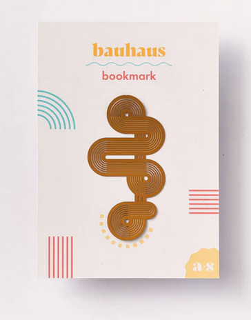 A brass-coated steel bookmark with a geometric abstract design, packaged on a card with the text 'bauhaus bookmark' and colorful geometric patterns.