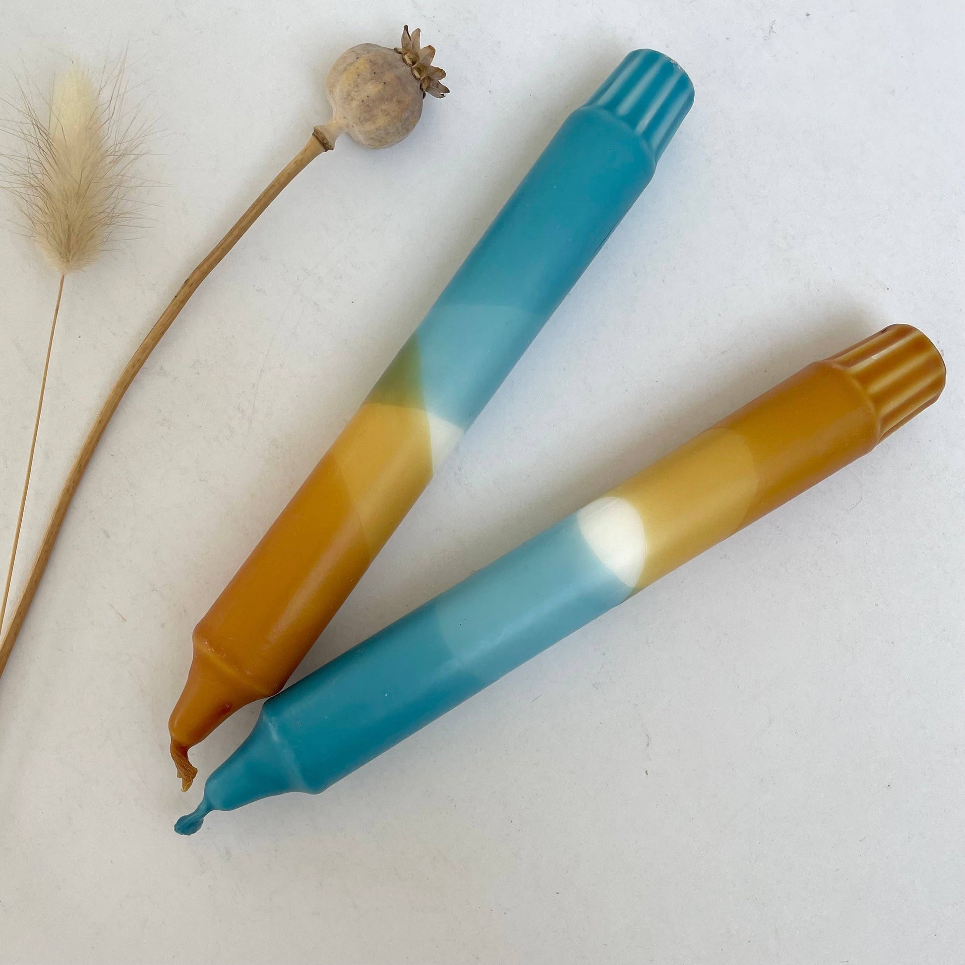 Two blue and caramel candles, on a white background with a dried plant. Hand dip dyed by The Singing Rabbit for Heart Gallery