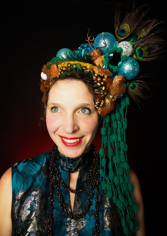 Lisa wearing a decorative headpiece with peacock feathers and beads against a black background for our Christmas Crowns and Conversations Event