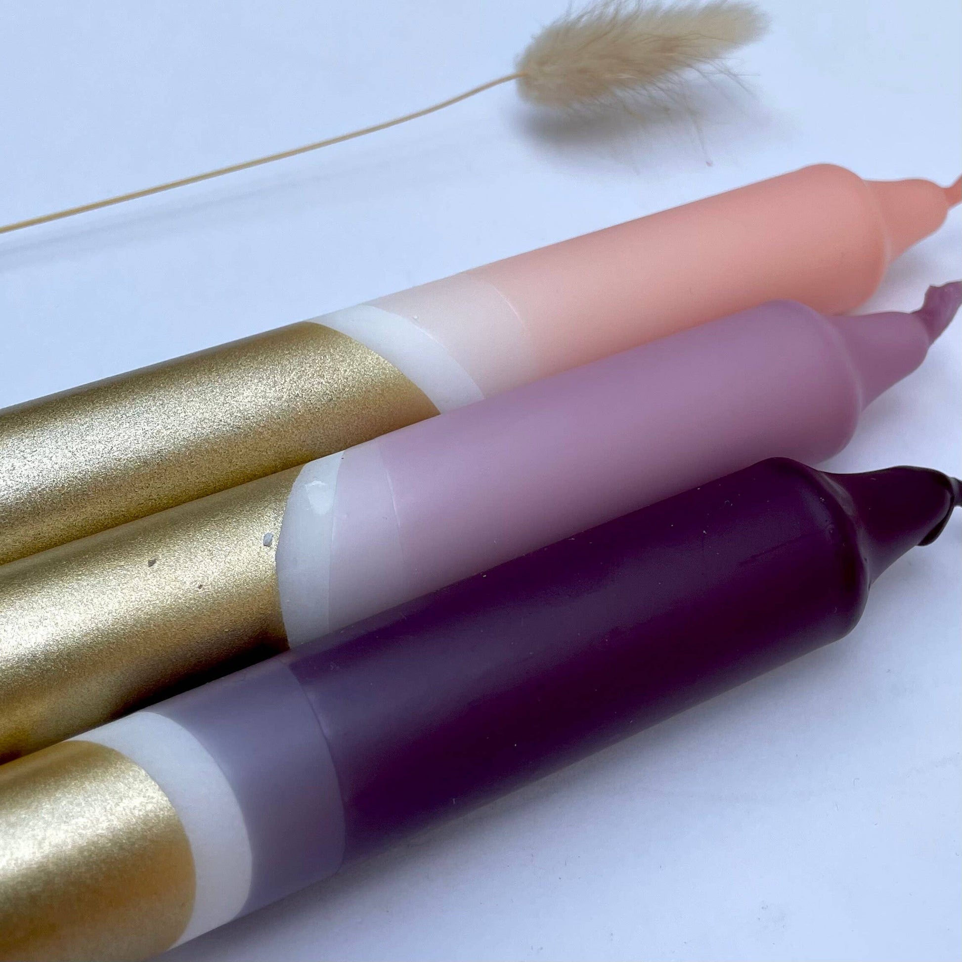 close up of a  set of three hand-dipped dinner candles in aubergine, heather, and blush pink with gold accents, displayed alongside a dried plant on a white background by The Singing Rabbit