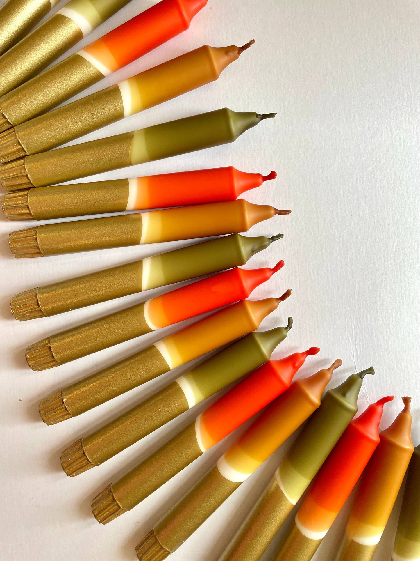A set of 3 dinner candles with a dip-dyed design, transitioning in colors from olive green to caramel, rust, and gold, arranged in a semi-circle on a light background.