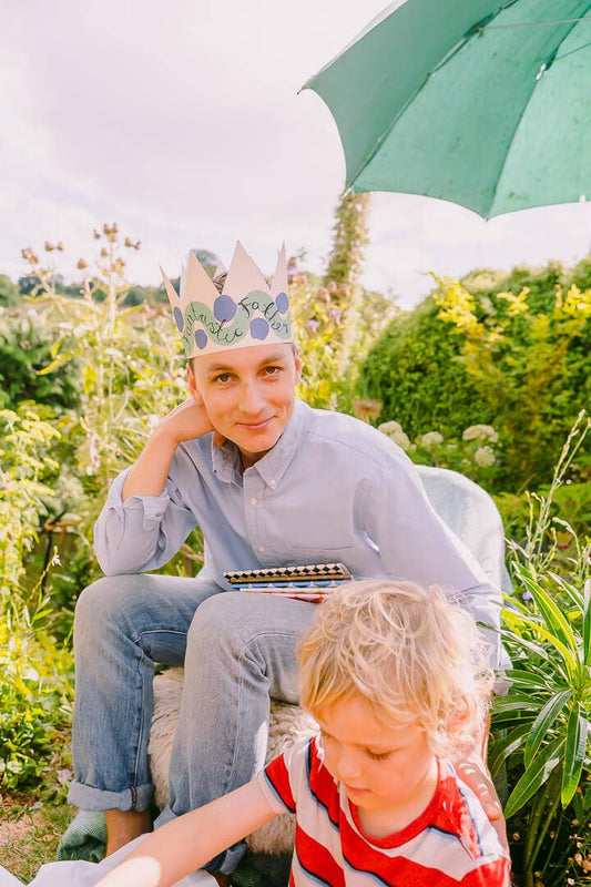 A Father wearing a paper crown, sitting on a rock with greenery around.