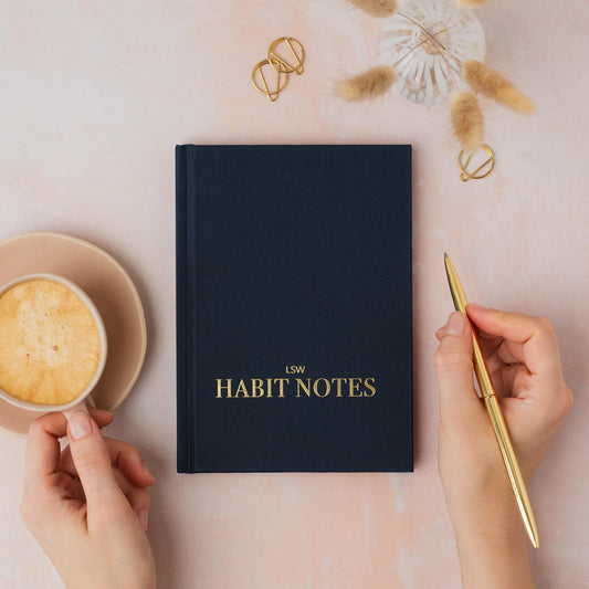Dark Blue notebook titled 'LSW Habit Notes' with a hand holding a gold pen and a cup of coffee on a light pink surface. LSW Habit Notes journal