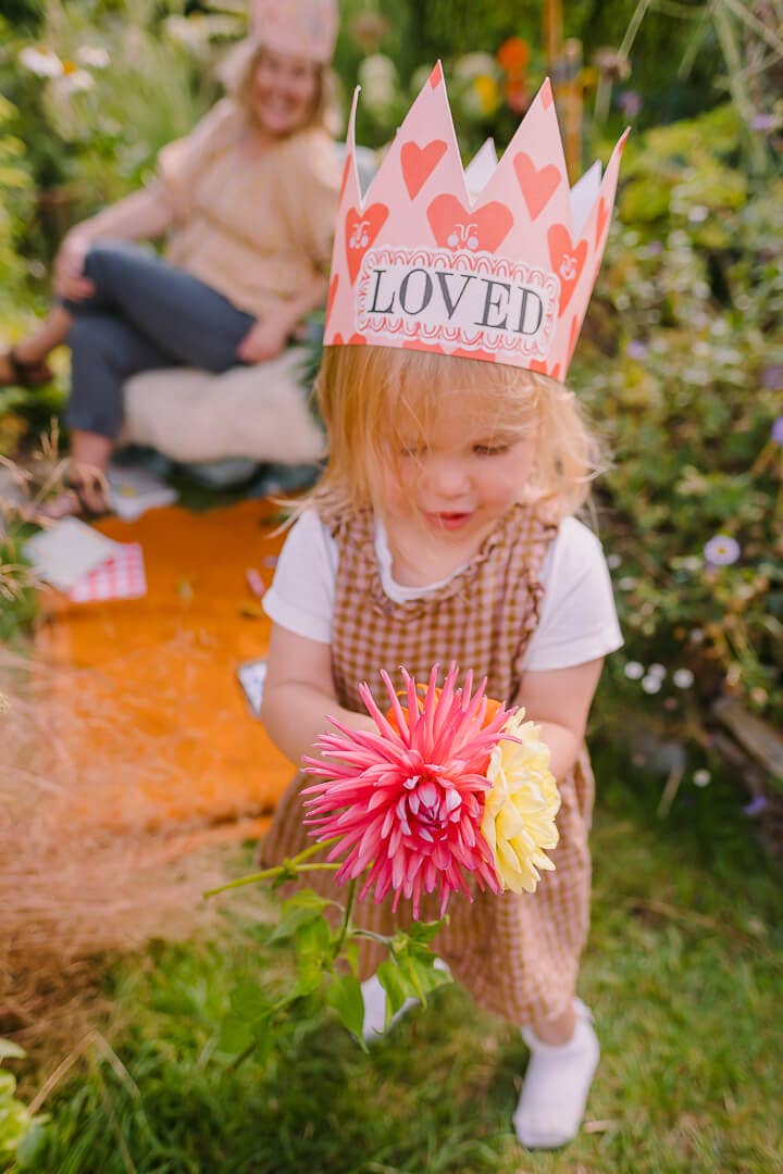 Child wearing a 'LOVED' crown holding flowers in a garden setting