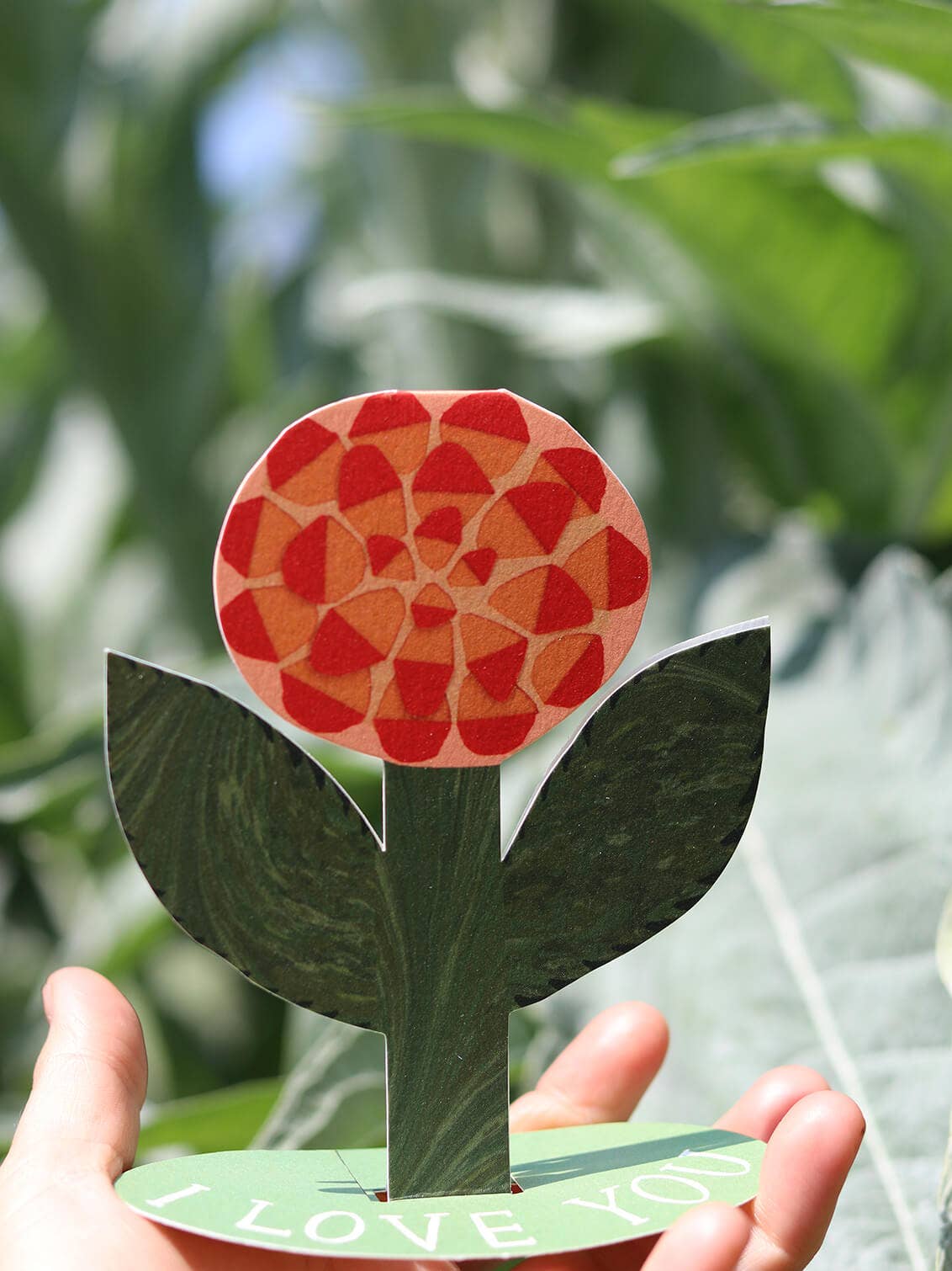 Cardboard flower with red pattern held by a hand against a green leafy background