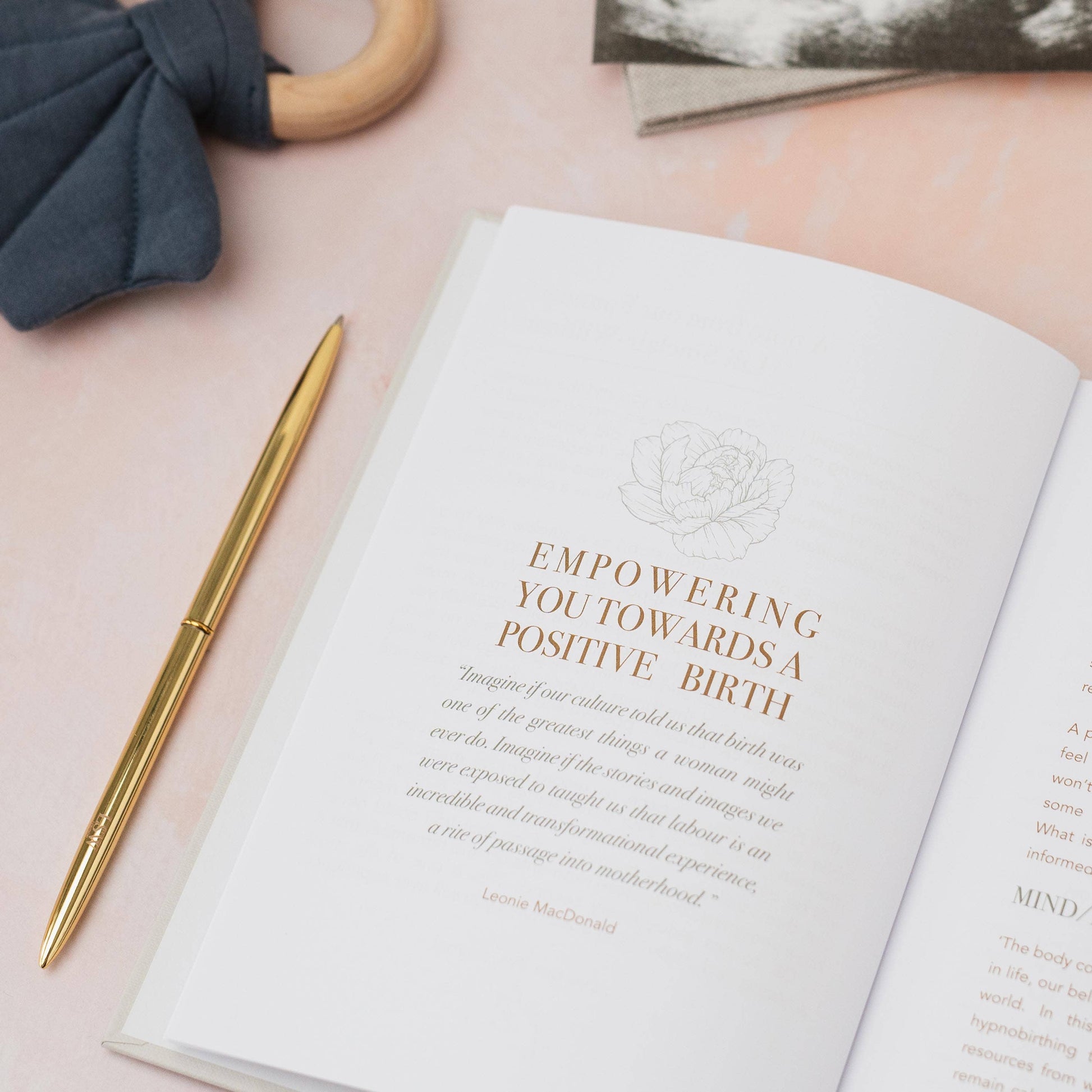 Open book with text on a pink surface next to a gold pen and gray fabric