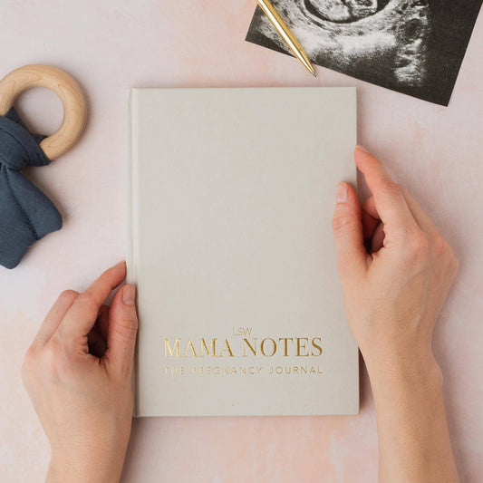 Pregnancy journal titled 'Mama Notes' held by hands with ultrasound photo and baby items on a light background
