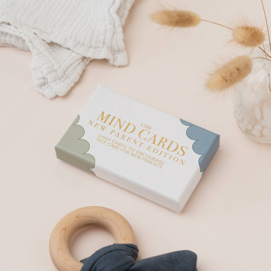 Box of 'MIND CARDS New Parent Edition' on a light surface with a wooden ring and textured fabric. LSW and Heart Gallery