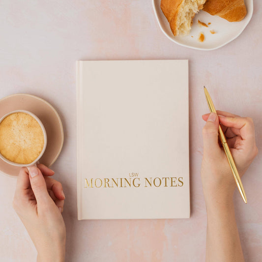 Notebook titled 'Morning Notes' with a hand holding a pen and another hand holding a coffee cup on a pink surface.
