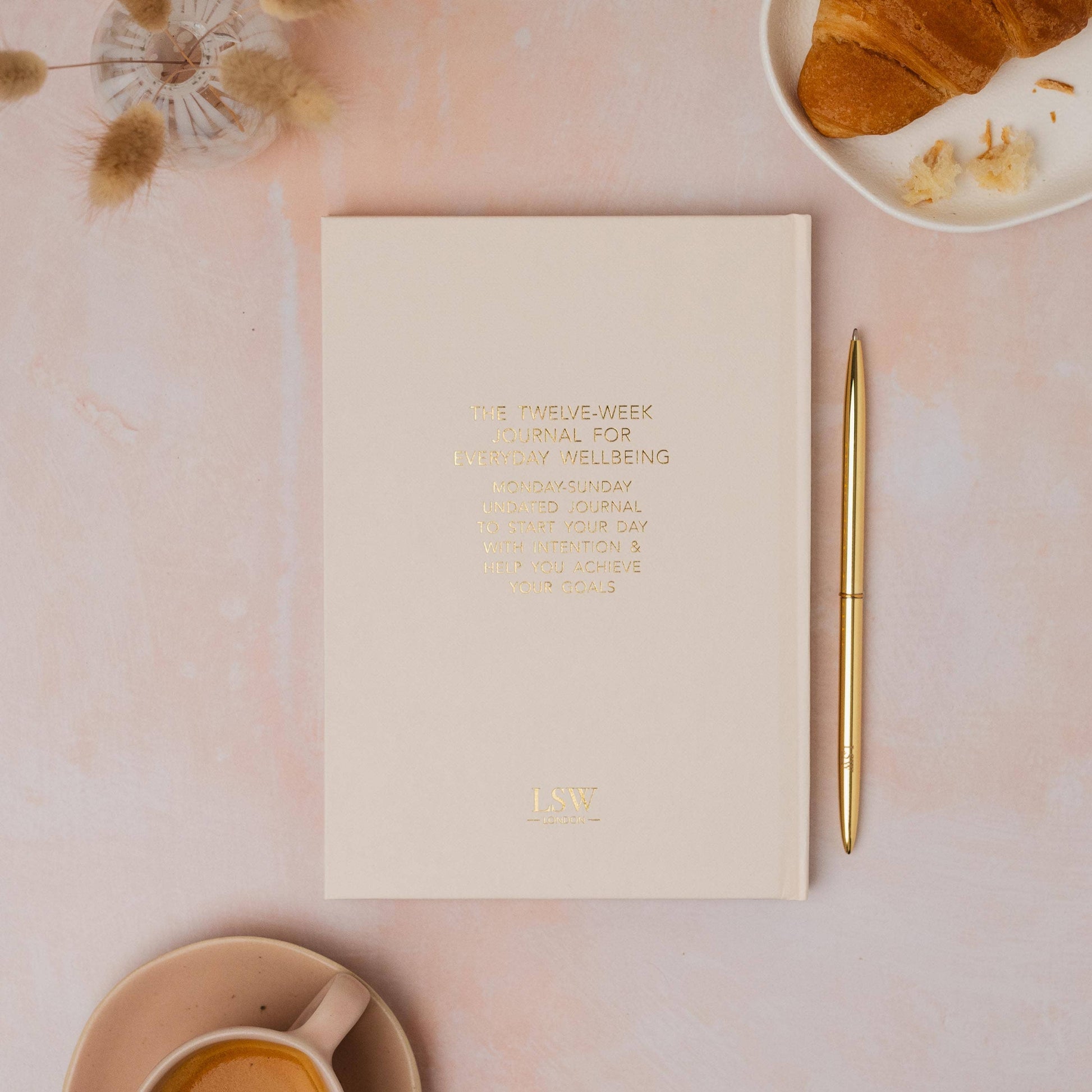 Notebook with a gold pen on a pink surface with a croissant and cup of tea.