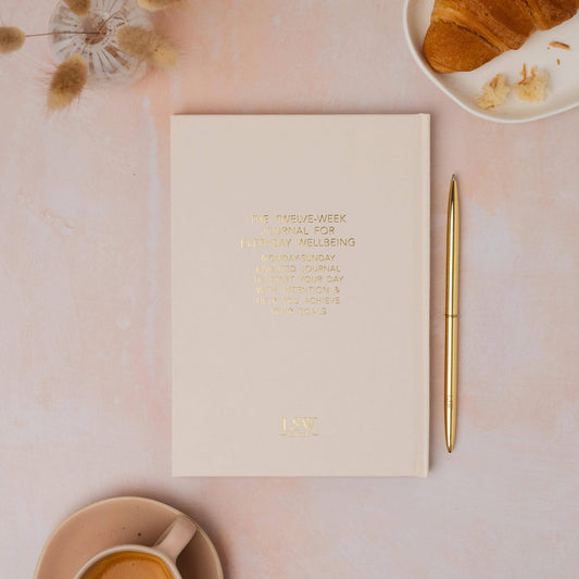Notebook with a gold pen on a pink surface with a croissant and cup of tea.