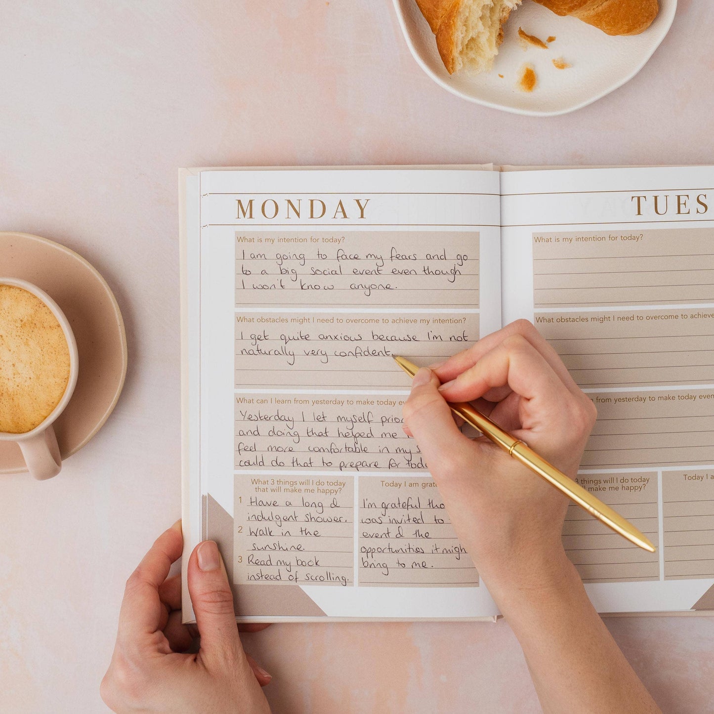 Person writing in a self help book with a cup of coffee and bread on a table.