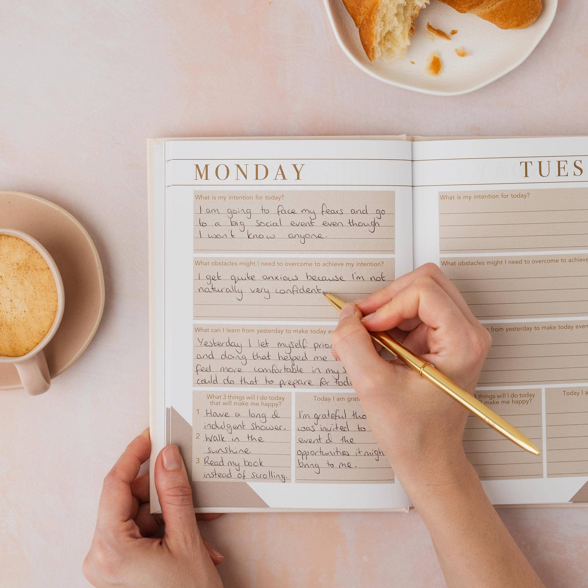 Person writing in a self help book with a cup of coffee and bread on a table.