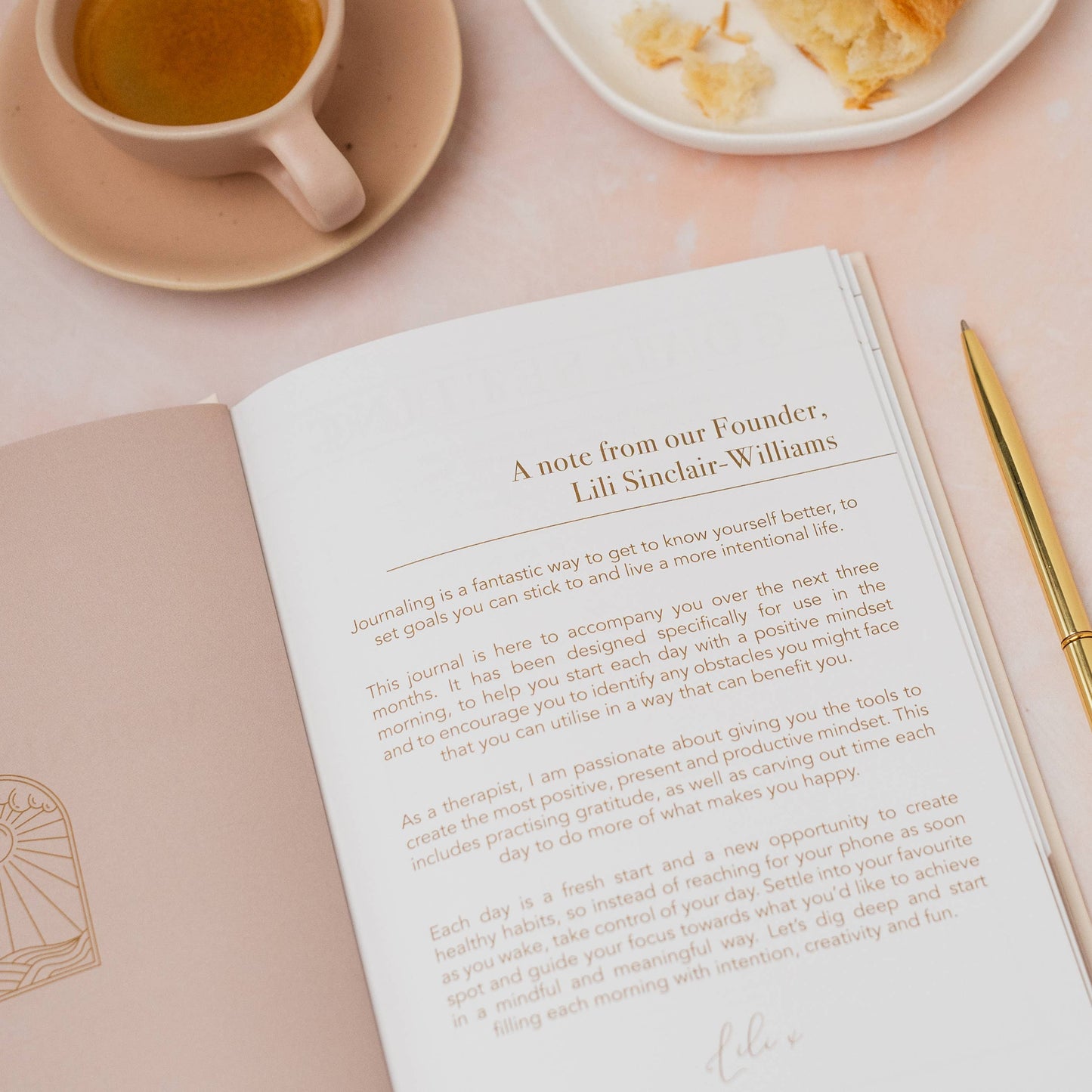 Open journal with a note from the founder, cup of tea, and pastry on a pink surface