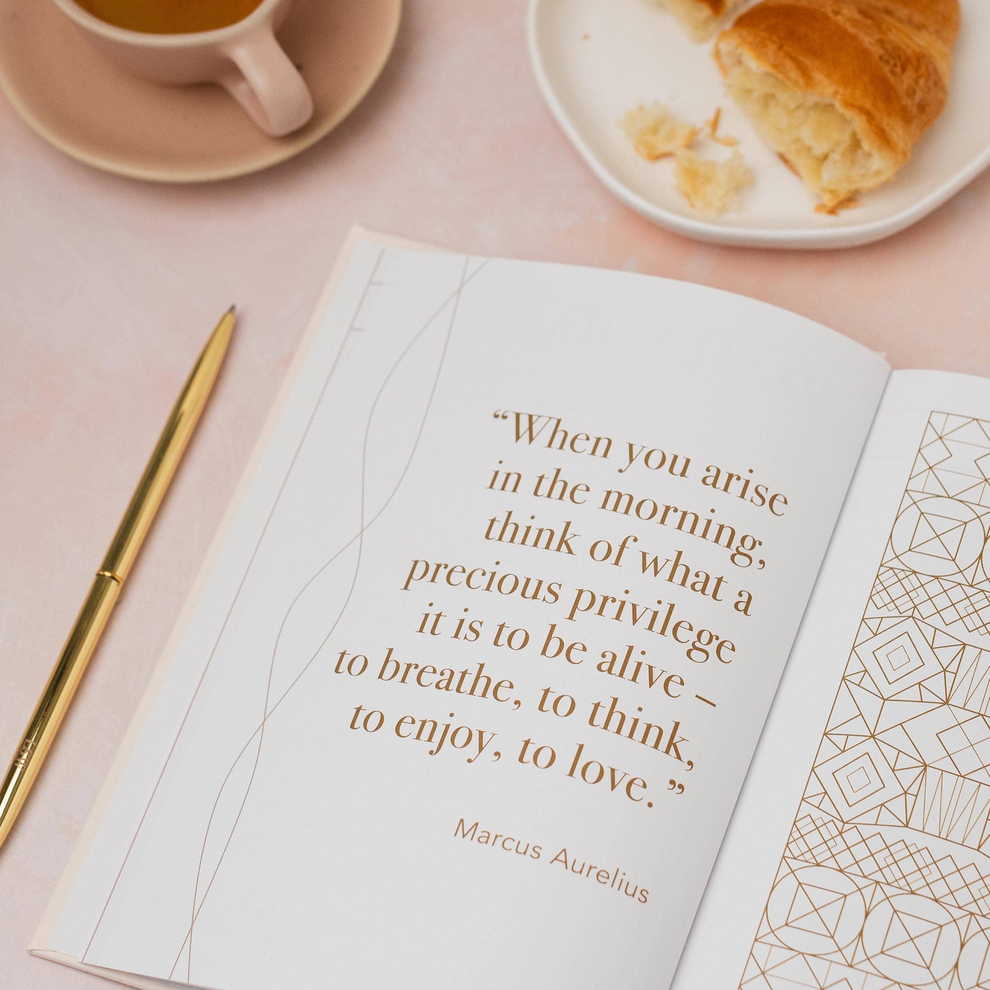 Open book with a quote by Marcus Aurelius, a cup of coffee, and a croissant on a pink surface.
