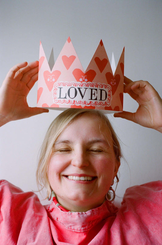 Person wearing a pink paper crown with hearts and the word 'LOVED' on a plain background