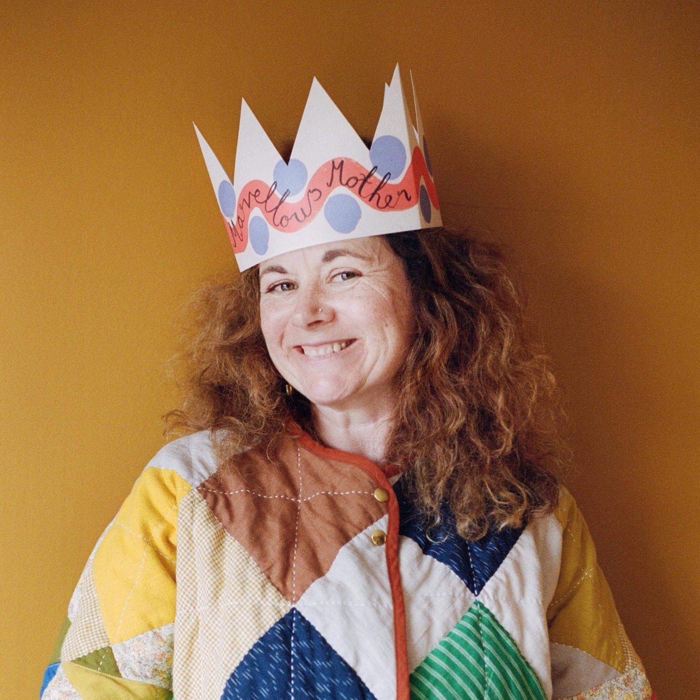 Person wearing a colorful paper crown with 'Well done Mother' text against a mustard yellow background