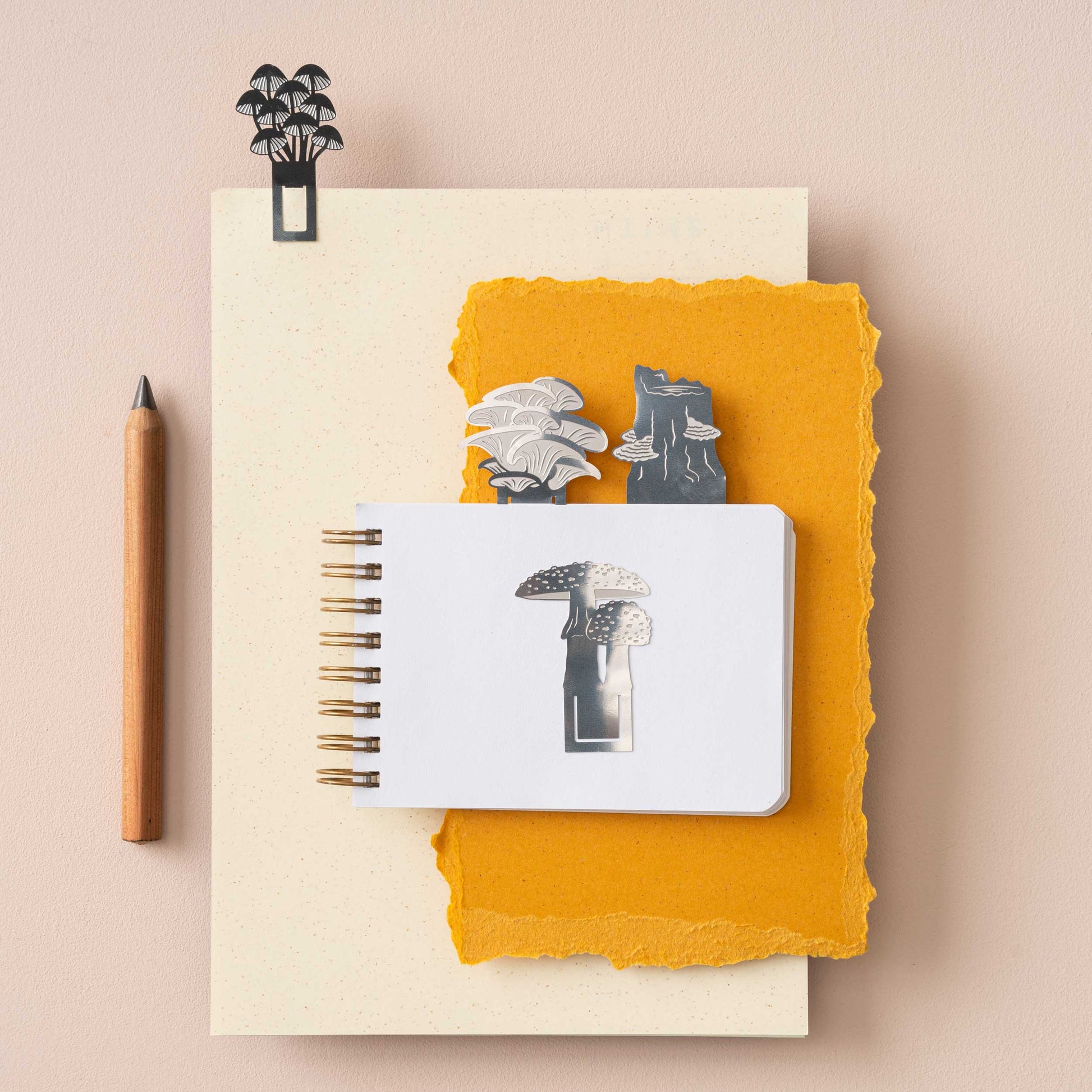 A set of stainless steel mushroom bookmarks in various designs, displayed on top of a notebook with a pen and decorative elements.