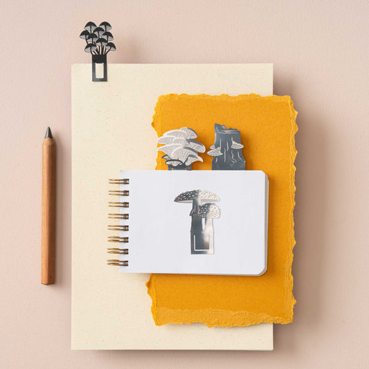 A set of stainless steel mushroom bookmarks in various designs, displayed on top of a notebook with a pen and decorative elements.