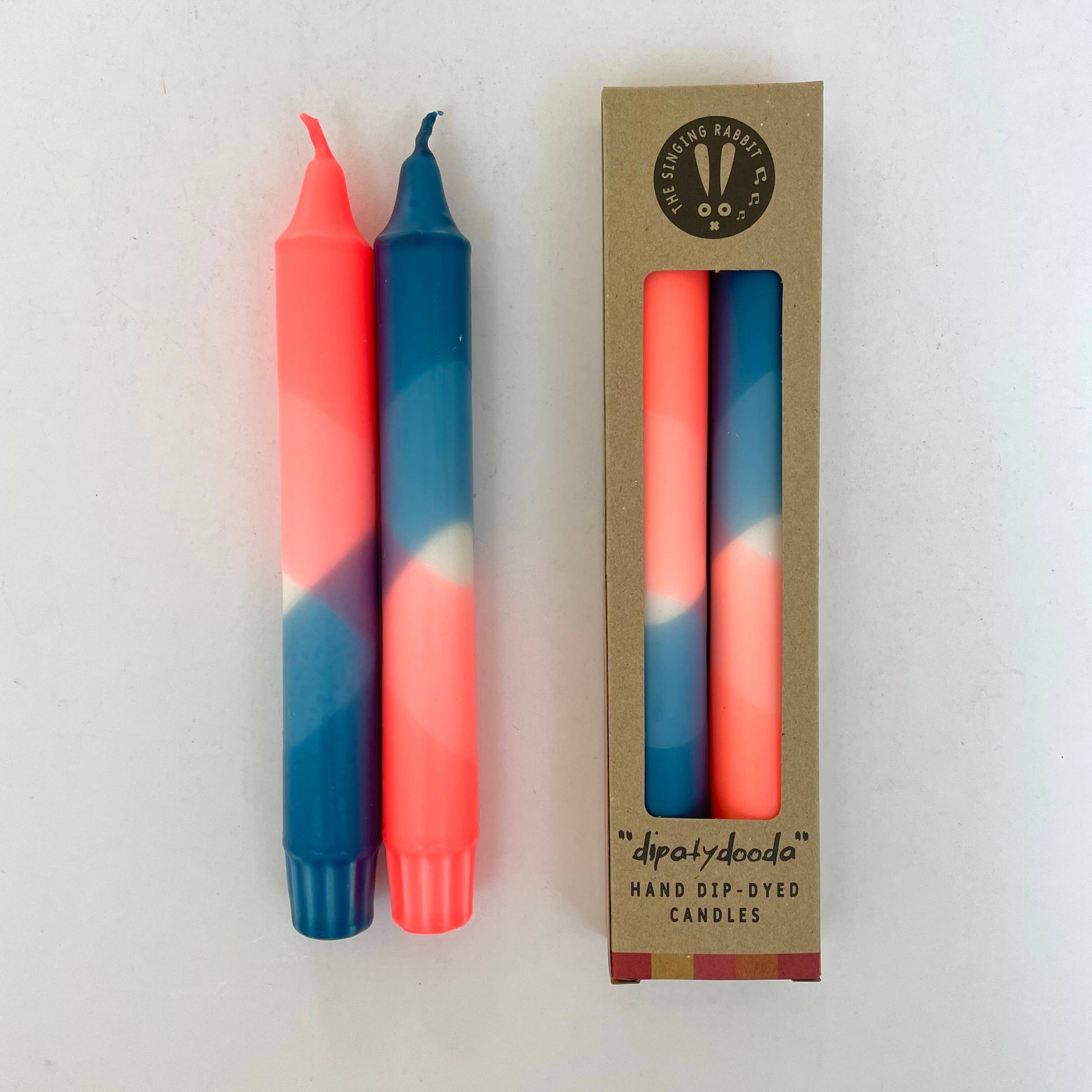 Two hand-dip-dyed candles in neon coral and teal blue with their packaging on a white background by The Singing Rabbit