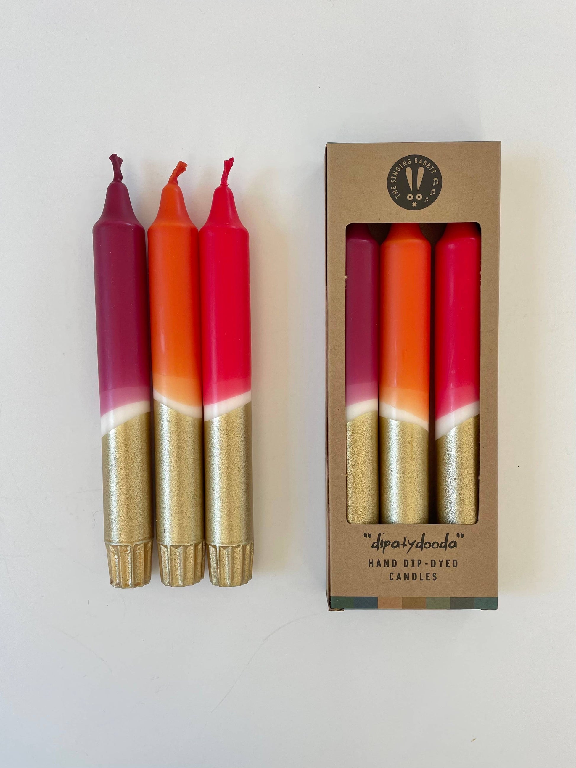 Three dip-dyed candles with gold and red gradient in front of their packaging box.