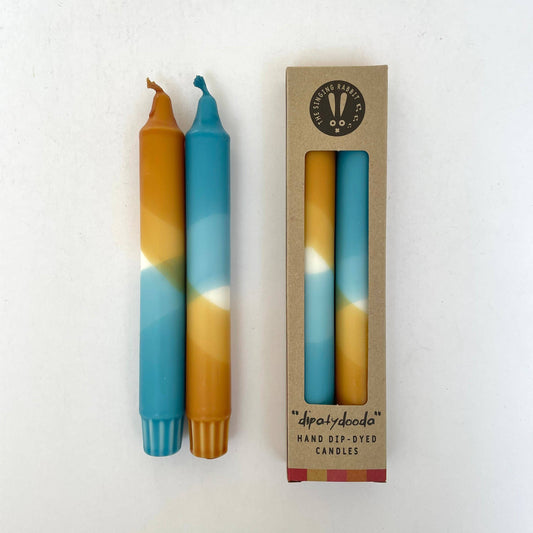 Two dip-dyed candles, blue and caramel, next to their packaging on a white background by The Singing Rabbit
