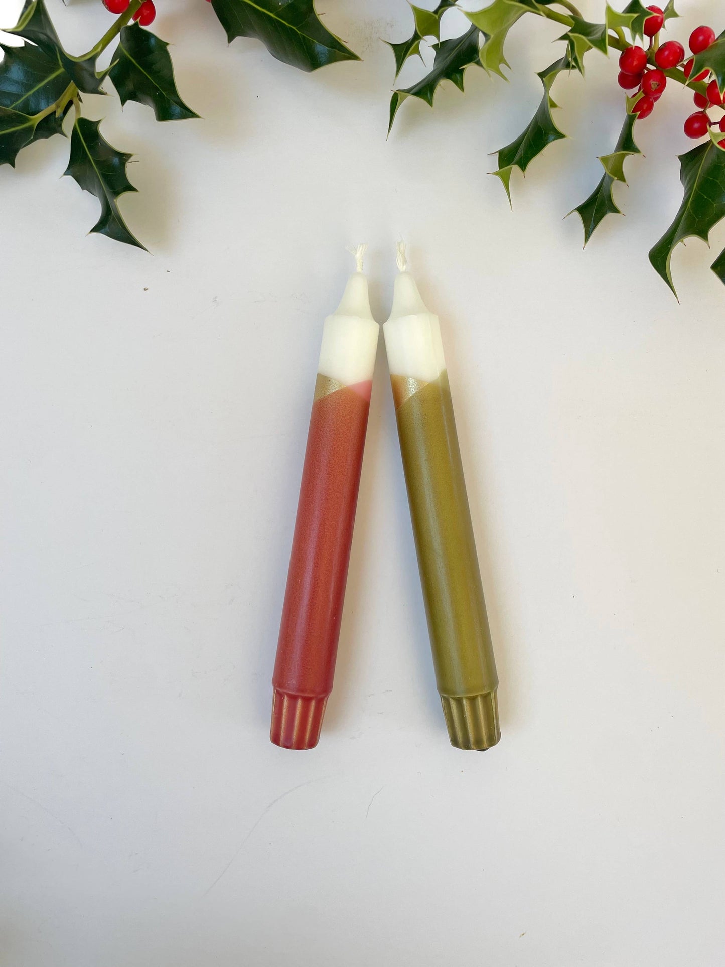 Two candles with red and green bases on a white surface with holly leaves and berries.