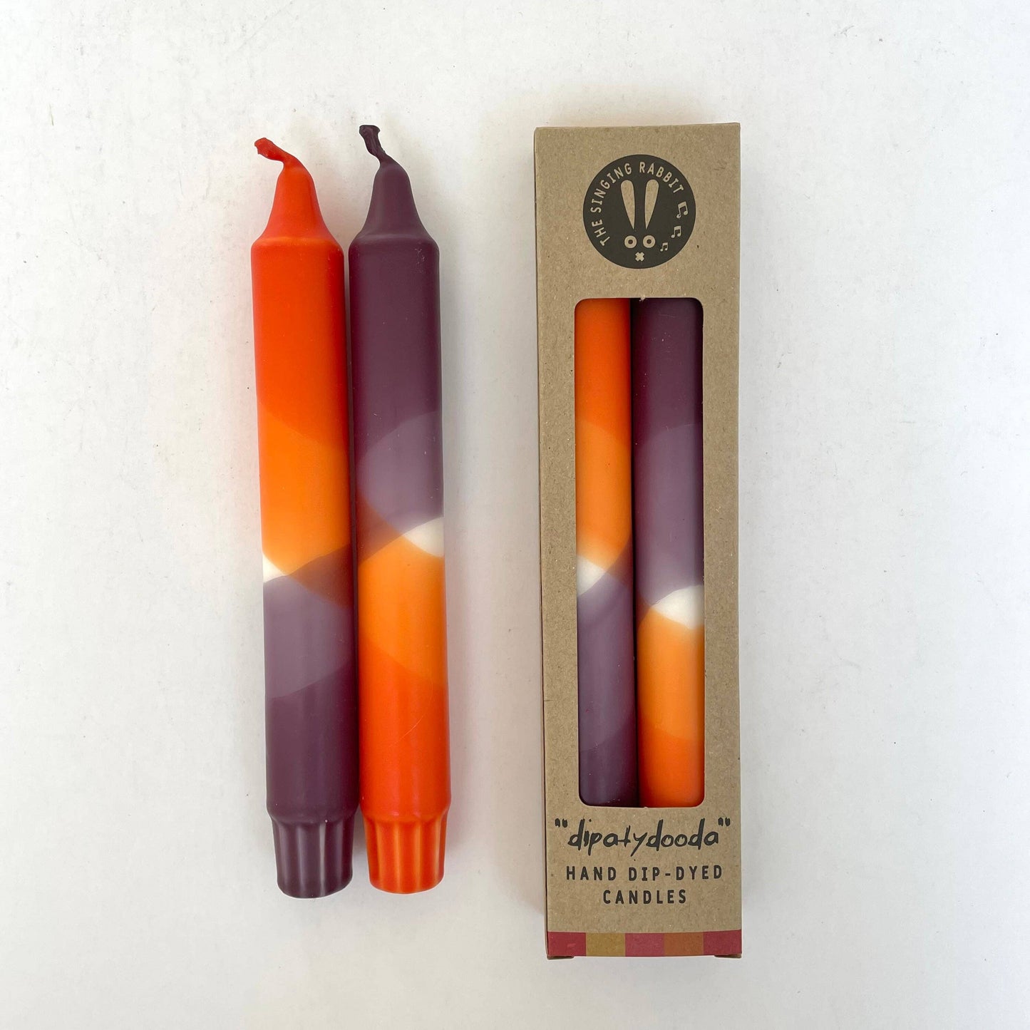 Two hand-dip-dyed candles with a gradient pattern and their packaging on a white background by The Singing Rabbit