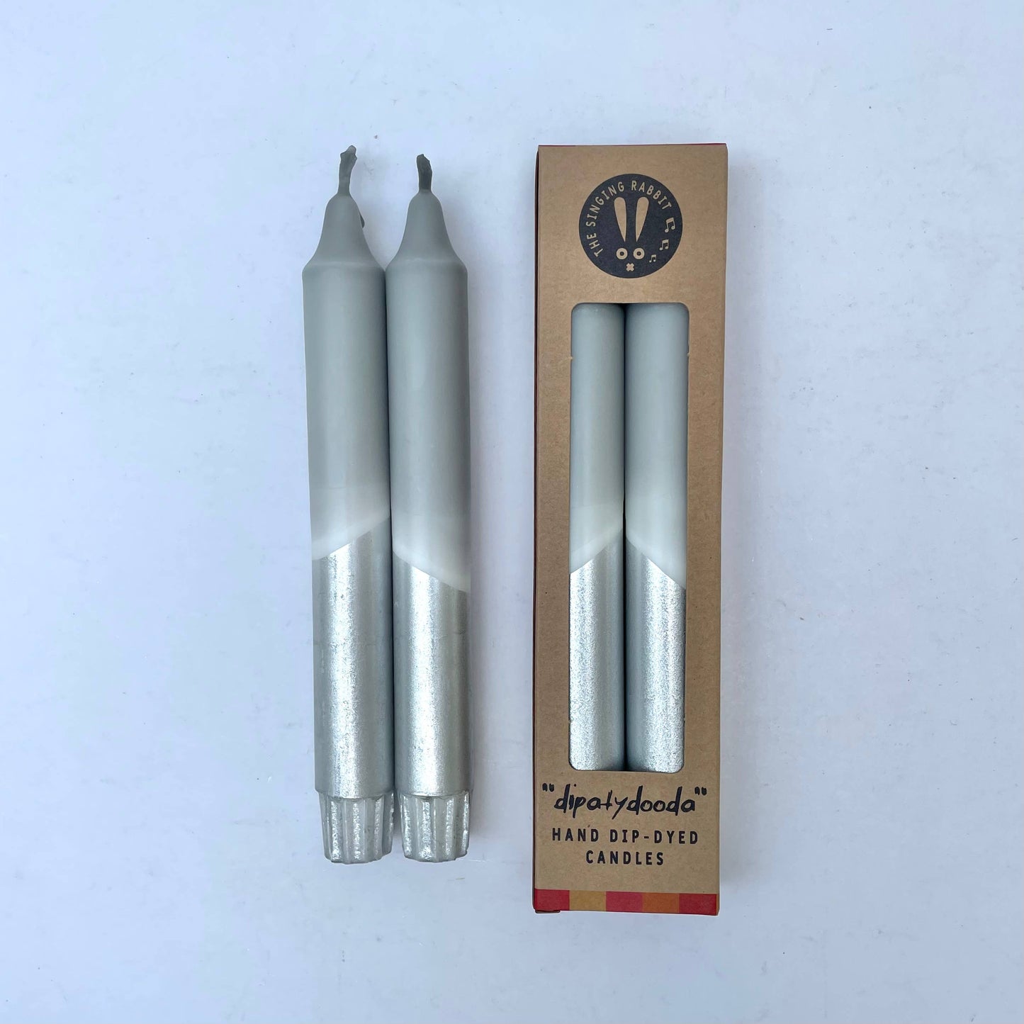 Two grey candles with silver bottoms next to their cardboard packaging on a white background